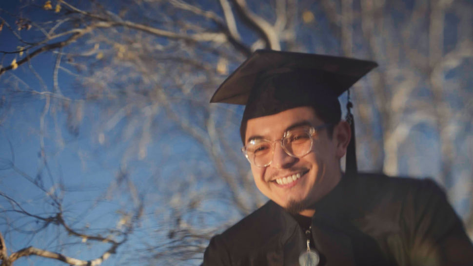 A smiling graduate in cap and gown stands outside under a clear blue sky, celebrating achievement.