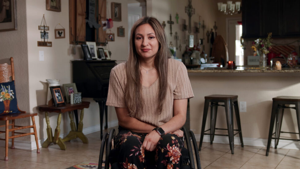 Woman in a wheelchair smiles while sitting in a cozy, decorated home with a kitchen and photo frames in the background.